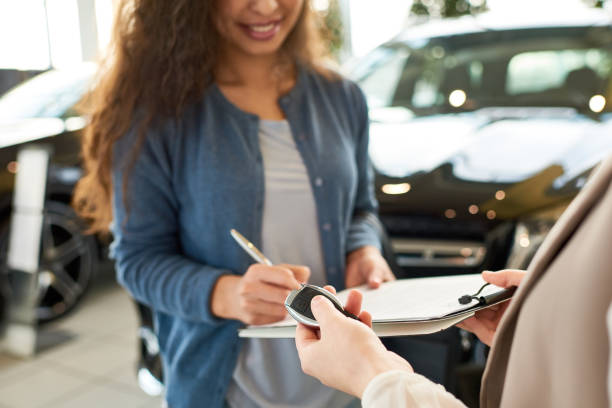 Signing Contract for New Car Close-up shot of smiling curly woman signing contract for new car, unrecognizable saleswoman holding key in hand, interior of modern showroom on background Car Hire stock pictures, royalty-free photos & images