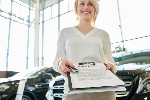 Low angle view of smiling young saleswoman in formalwear looking away while holding contract and pen in hands, interior of spacious car showroom on background
