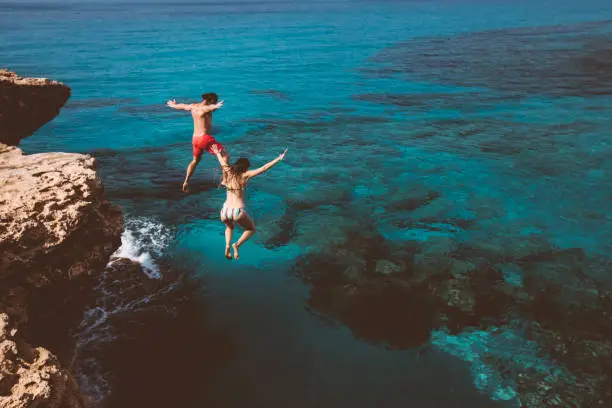 Young active man and woman diving from high cliff into tropical island blue sea water