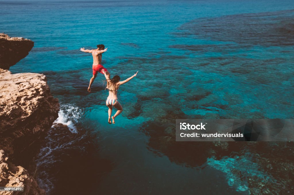 Young brave divers couple jumping off cliff into ocean Young active man and woman diving from high cliff into tropical island blue sea water Vacations Stock Photo