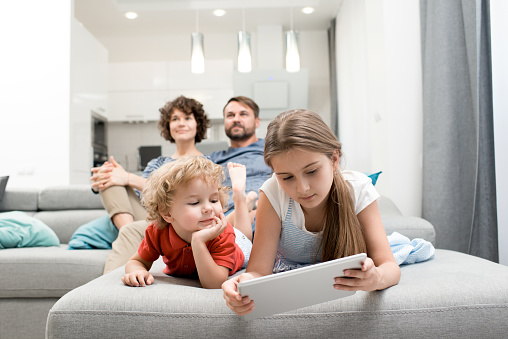 Cute little boy and his pretty elder sister lying on sofa and playing game on digital tablet while his parents sitting behind them and watching TV.