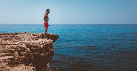 Young active diver standing on cliff looking at sea and preparing to dive into ocean