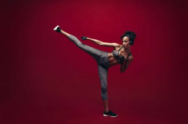 African female fighter practising high kick in studio. Full length shot of fit woman athlete performing a high kick over red background. Martial arts woman exercising taekwondo.