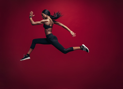 Female athlete running and jumping. Side view shot of healthy african woman working out against red background.