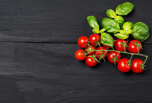 Ripe red cherry tomatoes with fresh green basil leaves on black wooden table, top view with copy space