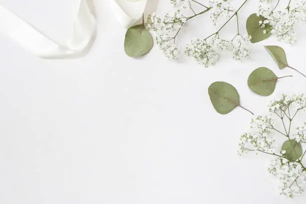 Photo of Styled stock photo. Feminine wedding desktop mockup with baby's breath Gypsophila flowers, dry green eucalyptus leaves, satin ribbon and white background. Empty space. Top view. Picture for blog