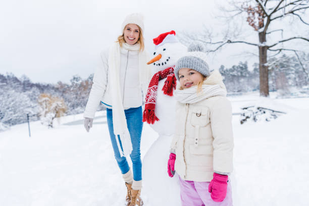 istock mother and daughter smiling at camera while standing with snowman 909343072