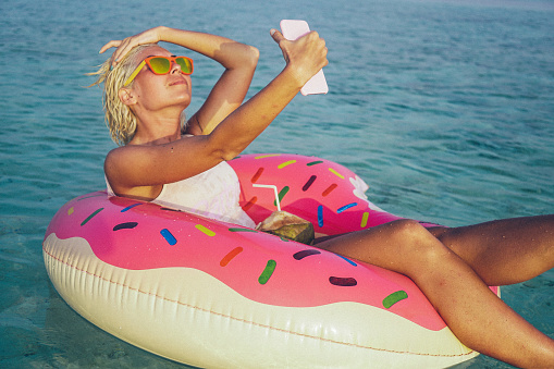 Young, attractive and beautiful woman is taking a selfie while floating in float in the ocean. Float is pink and yellow and have shape of doughnut witch is missing one part, something like one bite. She is wearing white swimsuit and orange sunglasses. In one hand she is holding smart phone while with other one she is fixing her hair. Coconut fruit is on her legs. Smile on her face is sign that she is relaxing and enjoying in this quite environment. Her short blonde hair is wet. Water is crystal clear and turquoise colored.