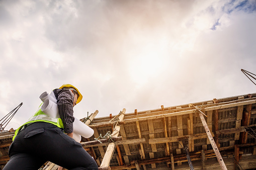 young professional engineer worker in protective helmet and blueprints paper on hand working on ladder at the house building construction site