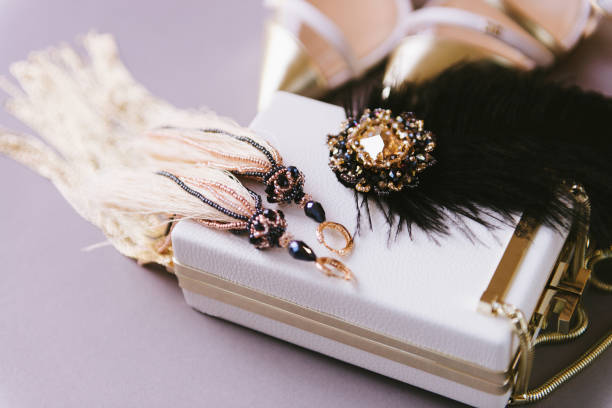Women's fashion accessories placed on lilac background. White leather handbag, gorgeous earrings and feathers brooch. Close up of modern lady essentials
