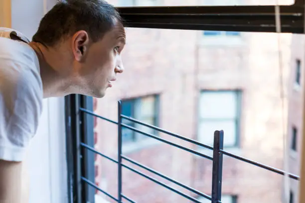 Photo of Closeup of young man's face looking outside of small apartment window in New York City NYC urban Bronx, Brooklyn brick housing, guard rail, security bars, checking weather