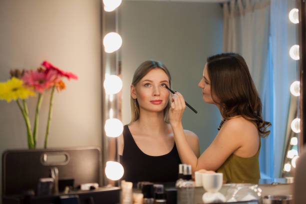 artista de maquillaje aplica maquillaje a atractiva chica rubia en el espejo con luces en el estudio de belleza. - 7676 fotografías e imágenes de stock