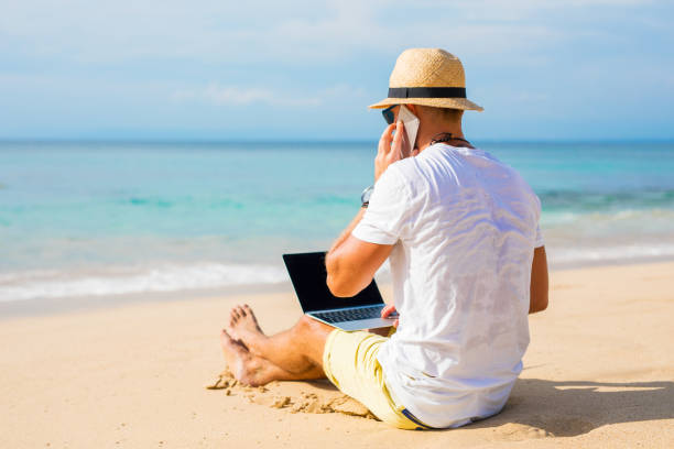 istock Young man on the beach working with laptop computer and speaking on the phone 909446358