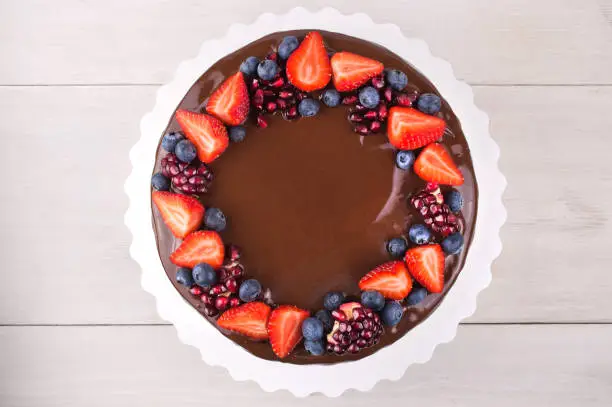 Birthday cake in chocolate with strawberries, blueberries and garnet on white wooden table. Top view. Picture for a menu or a confectionery catalog.