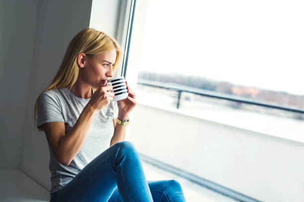istock Beautiful young woman drinking coffee and looking through window 909481698