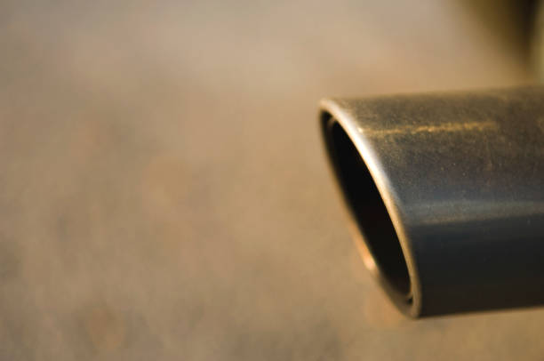 This color picture is of a Exhaust Pipe with Exhaust from a Car. the exhaust pipe is chrome silver and circular in shape. the exhaust pipe is connected to a vehicle. the lighting is sunlight reflecting off the metallic silver chrome tail pipe. 