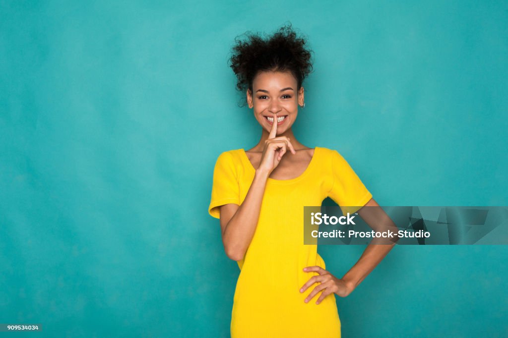 Secret. Young woman put finger on lips, hush sign Secret. Young african-american woman put finger on lips, hush sign, keep silence. Happy black girl on blue studio background Dress Stock Photo