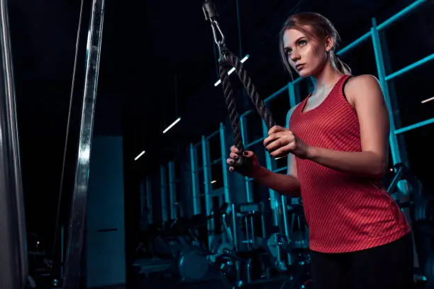 Beautiful girl doing exercises on the simulator in the gym dressed blank pink tank-top and tights. On a dark background the gym. Dressed in sport clothes.