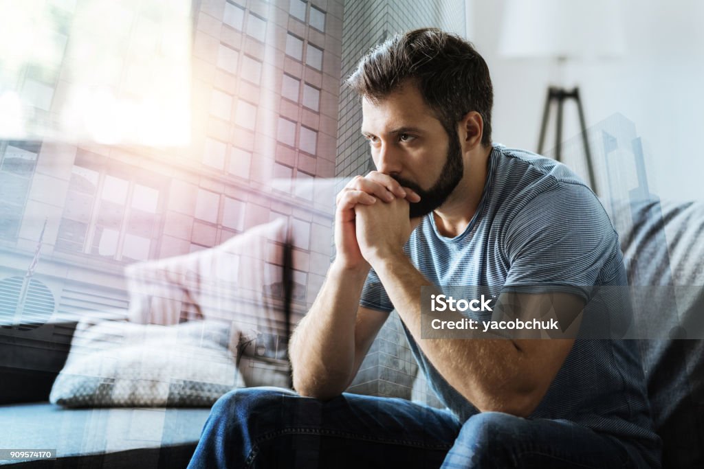 Thoughtful serious man sitting and thinking Think about it. Thoughtful serious beardful man sitting in the empty room holding hands near mouth and thinking. Men Stock Photo