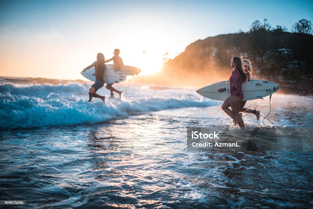 Friends running into the ocean with their surfboards Four friends, surfers, running into the water early in the morning with surfboards in their hands. Sun is rising in back. Surfing Stock Photo