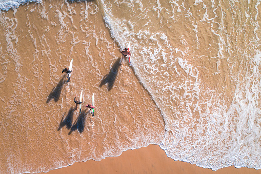 Aerial view of four surfers standing on the beach in Australia and going into the sea. They are holding surfboards in their hands.
