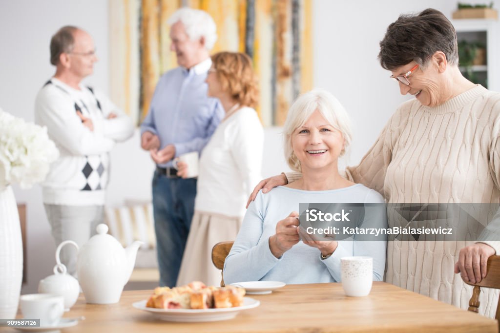 Elderly woman and smiling friend Elderly woman hugging smiling friend drinking tea during entertainment Retirement Community Stock Photo