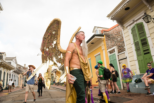 In New Orleans, USA a mature man wearing large golden angle wings walks along a French Quarter Street during Mardi Gras. People are visible walking in the background and standing in front of historic homes.