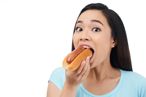 Head and shoulders portrait of funny Asian woman looking at camera with surprise while eating appetizing hot dog, isolated on white background
