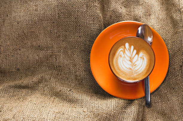 istock Top view hot latte in orange cup with floral pattern in foam on burlap background 909680776