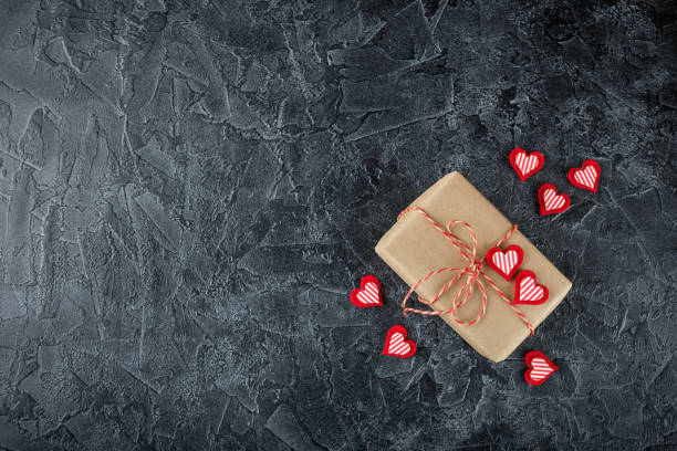 Vintage gift box (package) with blank gift tag on old wooden background. Gift boxes in kraft paper, decorative ribbon with wooden hearts and decorative felt hearts on a black background. Background for Valentine's Day. Flat lay. Top view. felt heart shape small red stock pictures, royalty-free photos & images