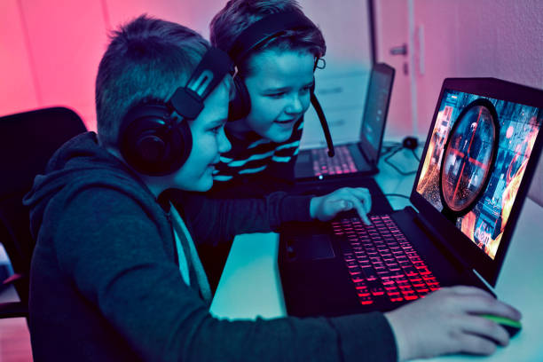Two boys sits in front of gaming laptops. They are brothers and both wear headsets and play a video game. It is night. 