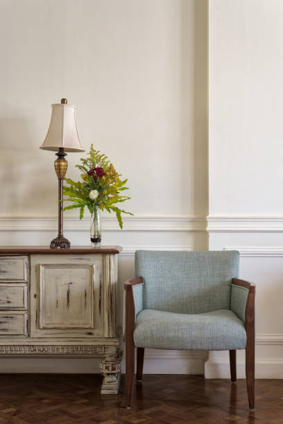 istock Light blue armchair and cream vintage sidebar with table lamp and flowers planter on off white wall and wooden floor 909718708