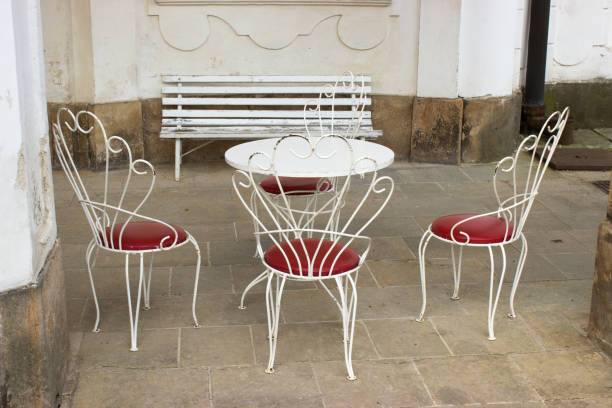 On the stone pavement are a metal chairs in the shape of a peacock with a red leather seat and round table. In the background there is  a flaked white bench.