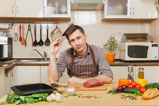 Tired sad caucasian young man in apron sitting at table with tablet, vegetables, cooking at home preparing meat stake from beef or pork, in light kitchen with wooden surface, full of fancy kitchenware