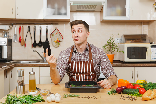 Perturbed stress young man in apron sitting at table with vegetables, talking on mobile phone, cooking at home preparing meat stake from pork, beef or lamb, in light kitchen with wooden surface