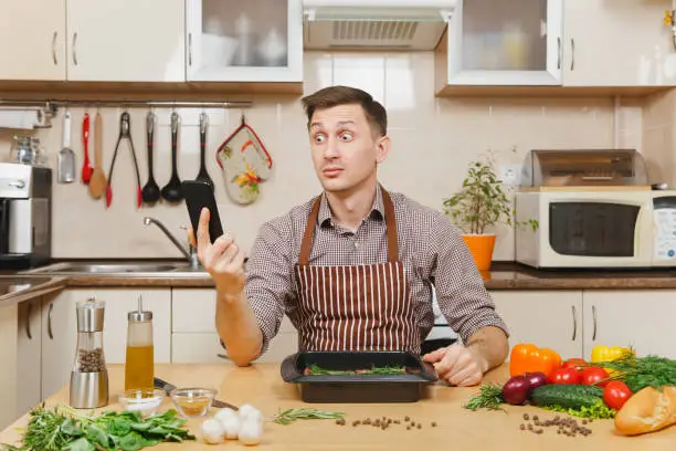 Photo of Perturbed stress young man in apron sitting at table with vegetables, talking on mobile phone, cooking at home preparing meat stake from pork, beef or lamb, in light kitchen with wooden surface.