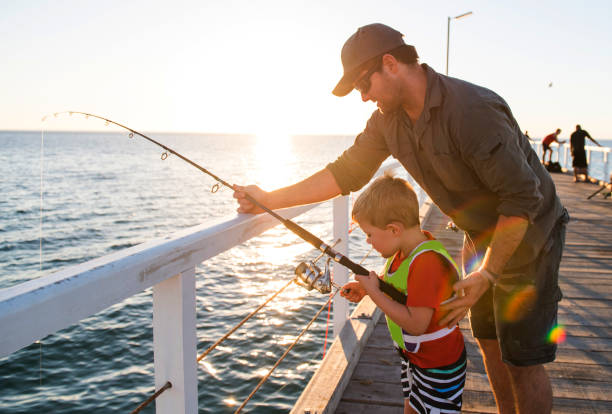 padre americano feliz enseñando a hijo poco joven para ser un pescador, a la pesca en mar muelle terraplén disfrutando y aprendiendo con la varilla de pescado en un día soleado de verano - embarcadero fotografías e imágenes de stock