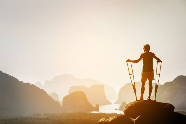 Disabled man with crutches on big rock stands like winner Disabled man with crutches stands on a big rock and looking at great view to islands and mountains. Space for text. Winner pose disabled person hiking outdoors stock pictures, royalty-free photos & images