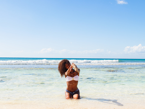 Scene of a young woman sitting in shallow water of the Caribbean sea and playing with sand. Behind her is a scenic view of blue sea against sky. Costa Romantica beach on Saona Island.