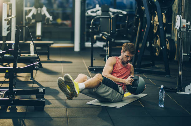 deportiva hombre haciendo un esfuerzo durante el ejercicio de abdominales con la bola de medicina en un gimnasio. - sit ups fotografías e imágenes de stock