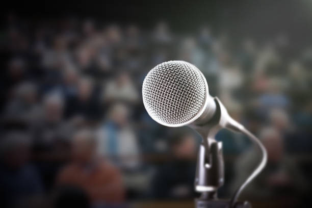 istock Brightly lit microphone with dark, defocussed audience in background 909908982