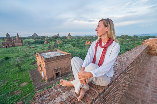 Caucasian young woman contemplating the Bagan archeological zone at sunrise, temples pagodas and monastery coming out from the lush greenery. People travel concept.