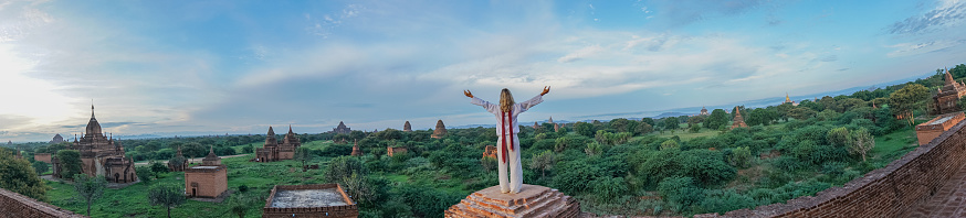 Caucasian young woman arms outstretched contemplating the Bagan archeological zone at sunrise, temples pagodas and monastery coming out from the lush greenery. People travel concept.