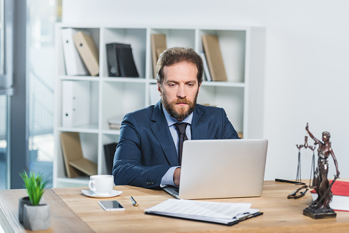 portrait of concentrated lawyer working on laptop at workplace with documents in office