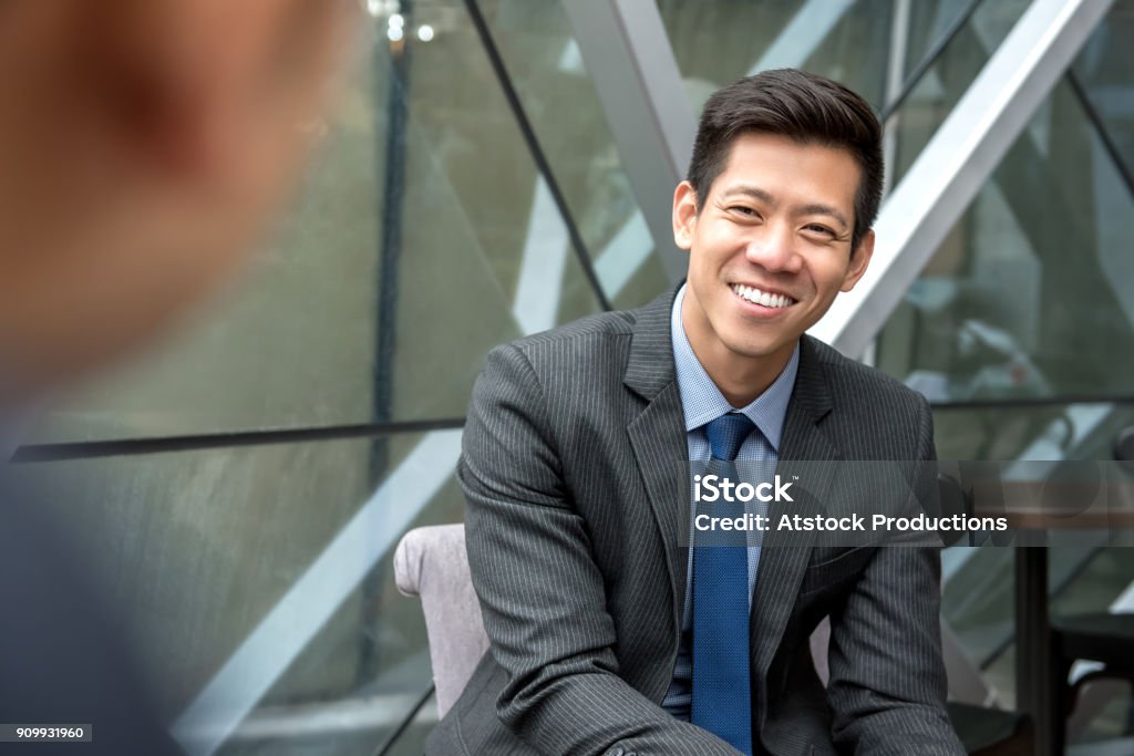 Asian businessman sitting at office lounge Friendly smiling handsome Asian businessman sitting at office lounge talking with his colleague Asian and Indian Ethnicities Stock Photo