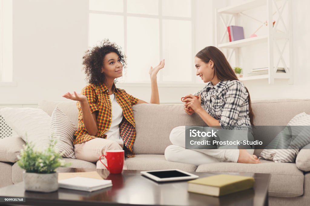 Two young female friends with coffee conversing Two happy young female friends with coffee cups conversing in living room at home, chatting about their life and relations, gossip and slumber party concept, copy space Friendship Stock Photo