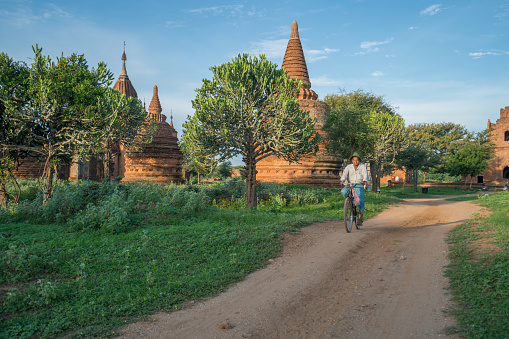 Burmese rural man driving bicycle on dusty road. Rural landscape in Bagan archeological zone, ancient buddhist temples in lushly nature in Burma countryside, Myanmar, Southeast Asia