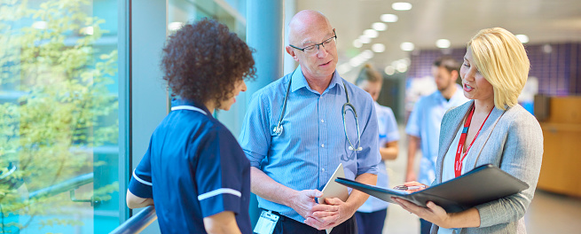 a business woman chats to a doctor and senior staff nurse in a busy hospital corridor