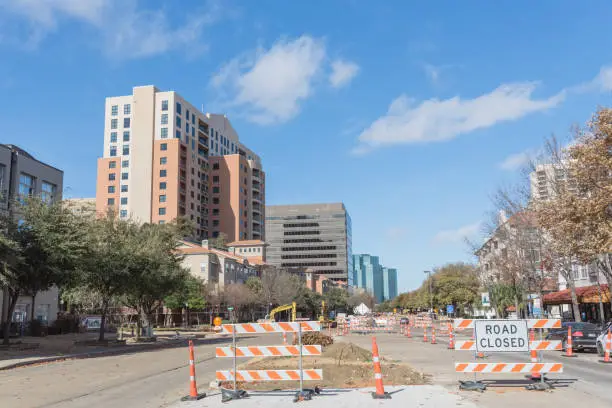 Road closed sign in Downtown Irving, Texas, USA under cloud blue sky. Barricade closures, cones with construction equipments and high-rise building in background.