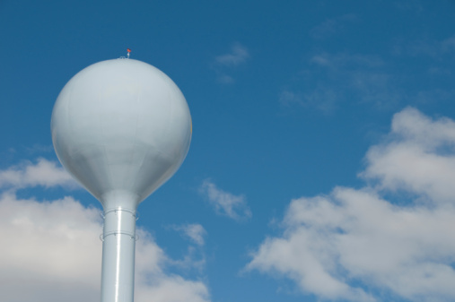 This image shows a low angle view of a red and white painted round shape water tower that resembles a fishing bobber, with view of tree leaves and blue sky.